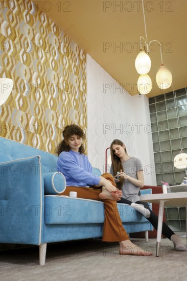 A woman is enjoying a pedicure while seated on a vibrant blue couch at home and chatting with her roommate