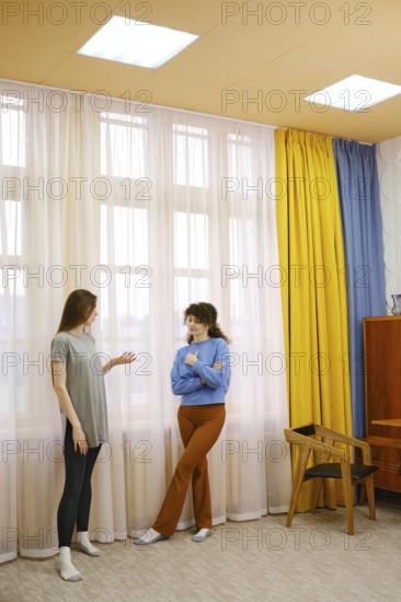Two women are talking in a bright room featuring large windows with flowing curtains in yellow and white