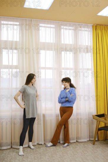 Two young women engage in a lively conversation in a well-lit room with large windows. The soft curtains filter sunlight, creating an inviting atmosphere for their friendly exchange