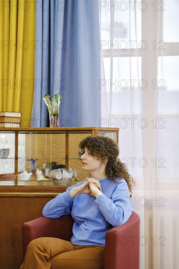 A woman with curly hair sits comfortably in a red chair, wearing a blue sweater and brown pants