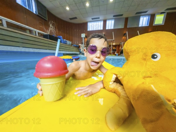 Boy with sunglasses sticking out his tongue, holding a refreshing drink and a plush toy while floating on a yellow inflatable mattress in a pool