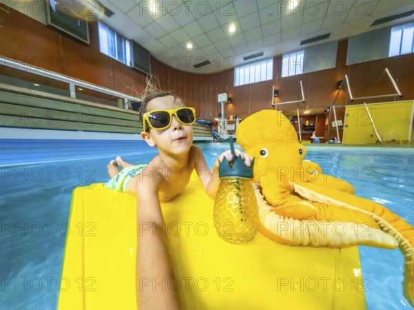 Young boy enjoys summer vacation, relaxing on a yellow float with a plush octopus and a refreshing drink in an indoor swimming pool