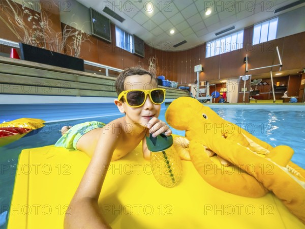 Young boy wearing sunglasses enjoying a refreshing drink while floating on a yellow raft with a plush octopus toy in an indoor swimming pool