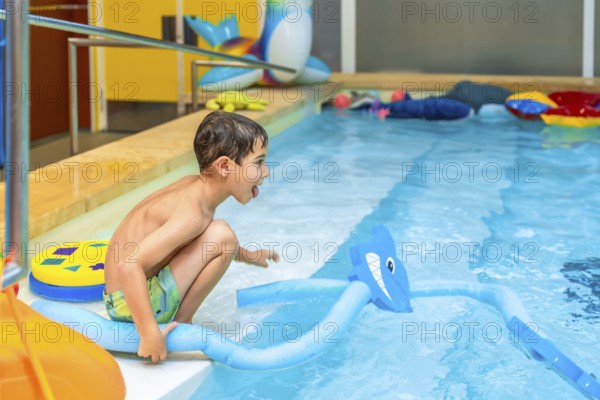 Happy child crouching at the edge of an indoor swimming pool, playing with a pool noodle shaped like a shark