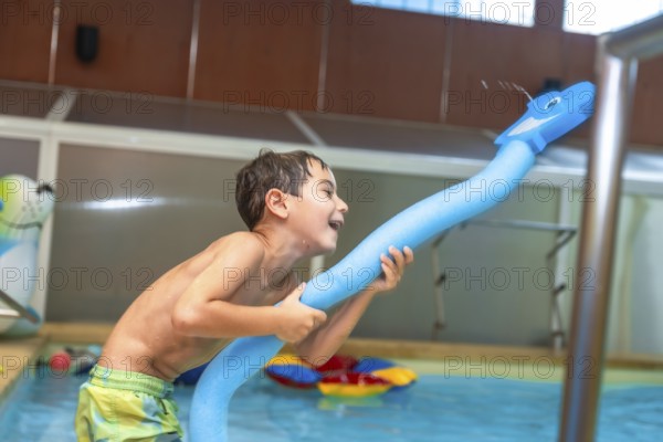 Happy child playing with a pool noodle shaped like a whale and spraying water in an indoor swimming pool