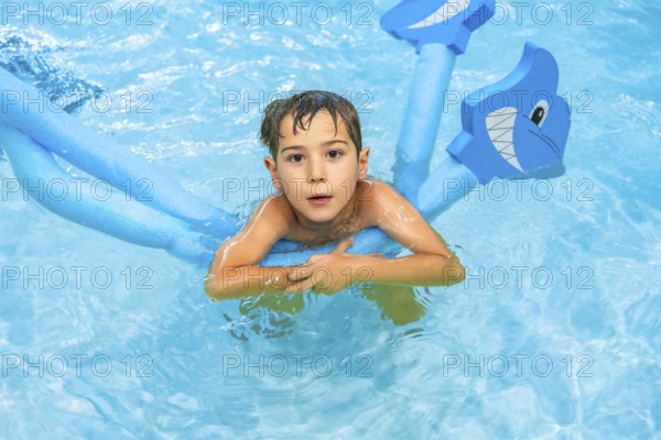 Young boy playing with a shark shaped pool noodle float in a blue swimming pool, enjoying his summer vacation