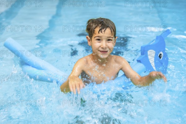 Smiling boy having fun in a swimming pool, playing with a pool noodle float shaped like a shark, enjoying summer vacation