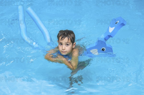 Boy swimming and playing with pool noodles and inflatable toy in a blue swimming pool during summer