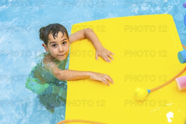 Young boy playing with a yellow floating mattress in a blue swimming pool during summer holidays
