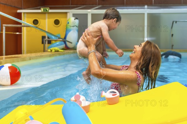Smiling mother lifting her baby while playing in a swimming pool during a sunny day, enjoying their vacation