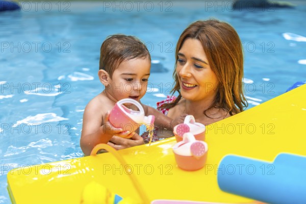 Mother and baby daughter enjoying time together, playing with pink and yellow toys while relaxing in a swimming pool during summer