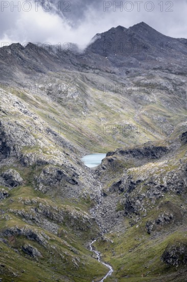 View of cloudy mountains and light blue mountain lake Trüber See in barren mountain landscape, Stubai Alps, near Ridnaun, South Tyrol, Italy