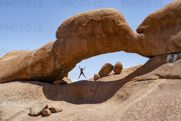 Tourist jumping in a rock arch, round rock formations with stone arch, Pontok Mountains, Great Spitzkoppe, Spitzkoppe, Great Spitzkoppe Nature Reserve, Namibia