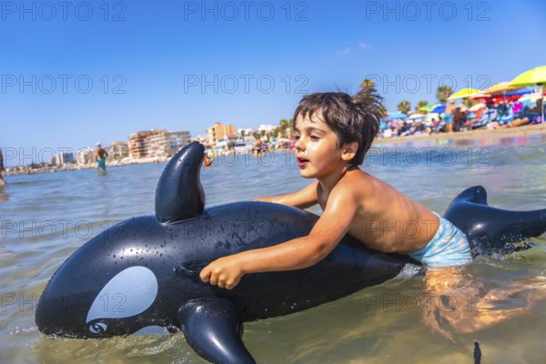 Happy child riding on an inflatable orca toy in the sea, splashing through waves and enjoying a joyful summer vacation by the beach