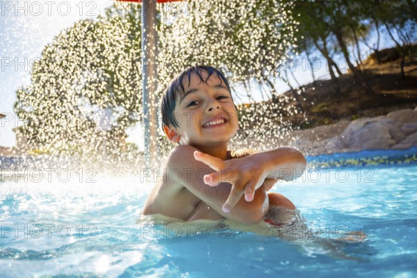 Smiling boy enjoying playful moments under a refreshing water shower in a swimming pool on a bright, sunny summer day