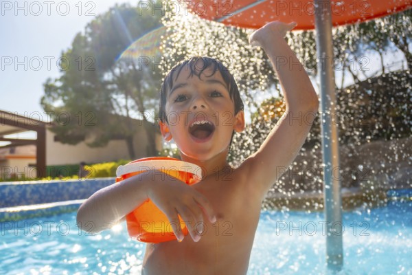 Boy holding orange bucket and raising arm under refreshing water shower in swimming pool on sunny summer day