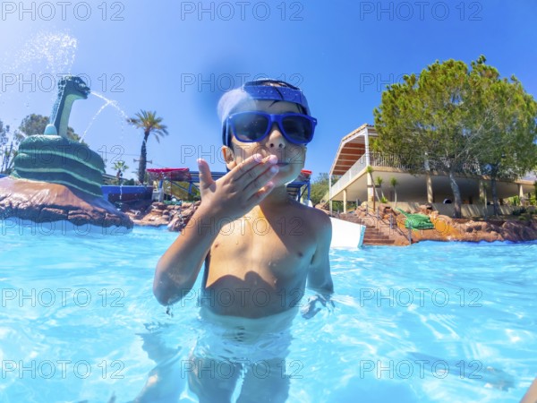 Young boy wearing sunglasses blowing a kiss while standing in a swimming pool at a water park with a snake shaped fountain in the background