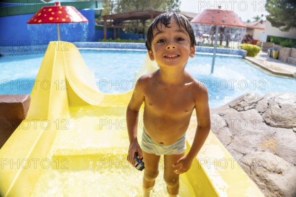 Smiling boy holding a toy car, enjoying summer vacation while sliding down a bright yellow waterslide at an exciting aqua park