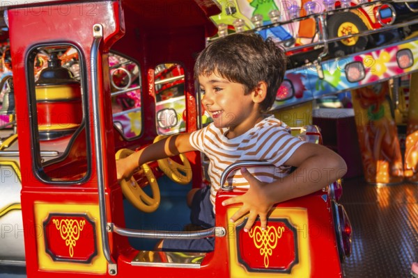 Young boy smiling while steering a vibrant red amusement park ride, surrounded by colorful decorations and lights, capturing the essence of childhood fun