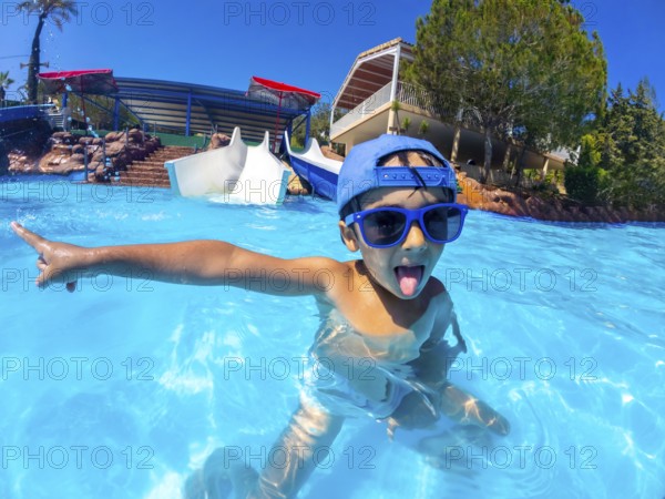 Playful child with blue sunglasses and hat pointing with finger and sticking tongue out in a swimming pool at a water park