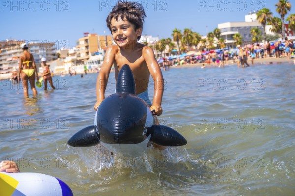 Smiling boy riding an inflatable orca toy in the sea waves, enjoying summer vacation at a crowded beach