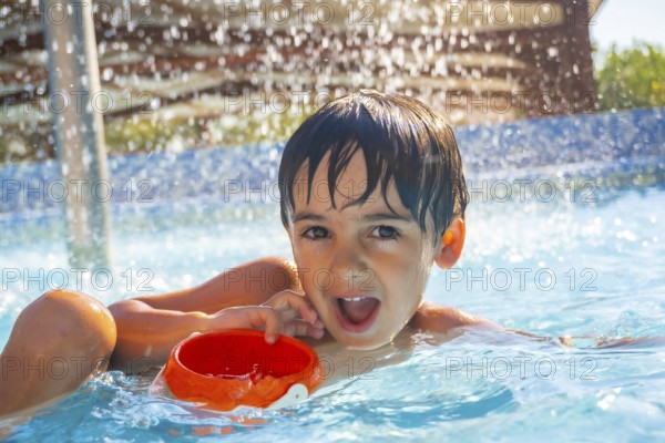 Portrait of a smiling child playing with an orange plastic bucket in a swimming pool under water jets on a summer sunny day