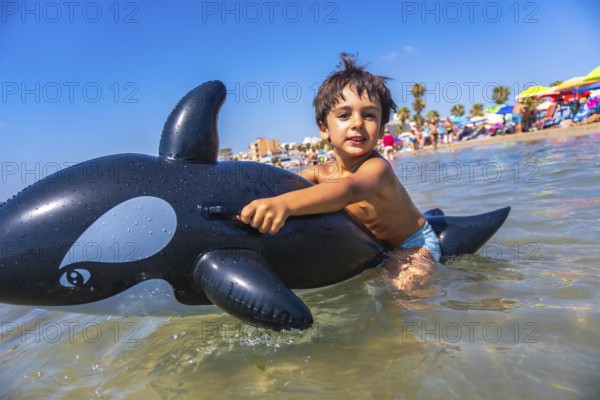 Young boy enjoying summer vacation, riding an inflatable orca toy in the ocean at a crowded beach