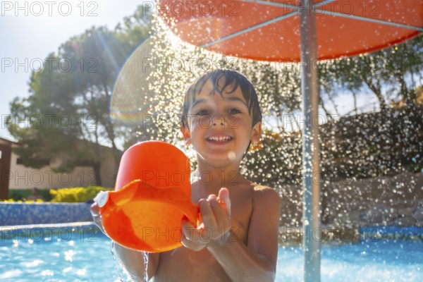 Smiling child enjoying a sunny day at the pool, holding an orange bucket under a refreshing water fountain, surrounded by trees and clear sky