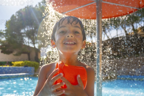 Smiling boy holding an orange water toy under a splash of water, enjoying a sunny day by the pool with trees in the background