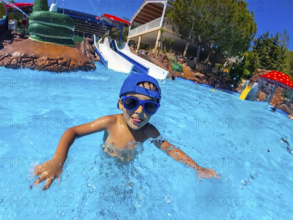 Young boy wearing sunglasses and a blue cap, enjoying a summer day at a water park, swimming in the pool