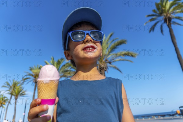Young boy wearing sunglasses and a cap enjoys a strawberry ice cream cone on a sunny beach with palm trees