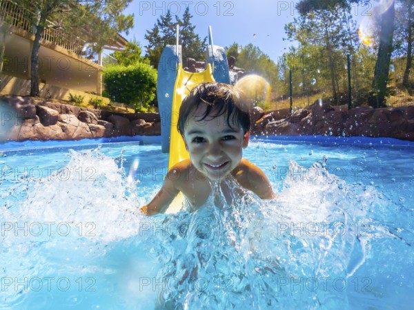 Young boy enjoying a sunny day, sliding into a vibrant blue pool with a big splash, surrounded by trees and clear skies
