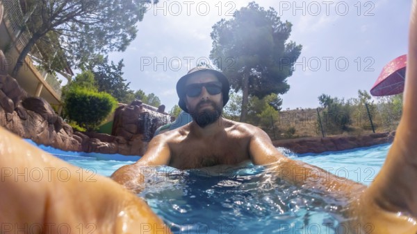 Bearded man wearing bucket hat and sunglasses enjoys relaxing in a water park pool on a sunny day, taking a selfie