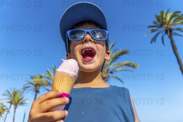 Young boy with sunglasses and cap enjoying a strawberry ice cream cone on a hot summer day with palm trees and blue sky