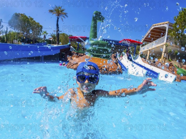 Happy child wearing blue baseball cap and swimming goggles, playing and splashing in a pool during summer holidays