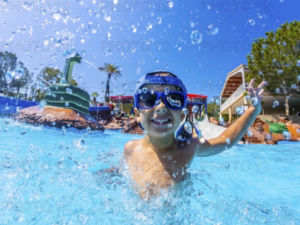 Happy boy wearing swimming goggles, splashing joyfully in a vibrant water park pool during summer vacations, enjoying every moment