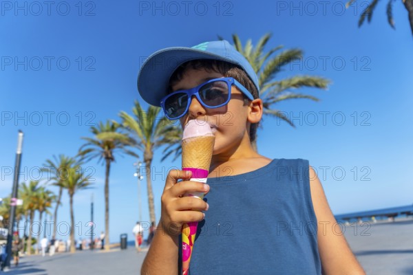 Young boy wearing sunglasses and cap enjoying a refreshing ice cream cone on a sunny summer day at the beach, with palm trees in the background