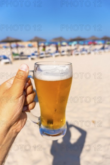 Hand holding glass of cold beer on beach with blurred beach umbrellas and sunbeds in background