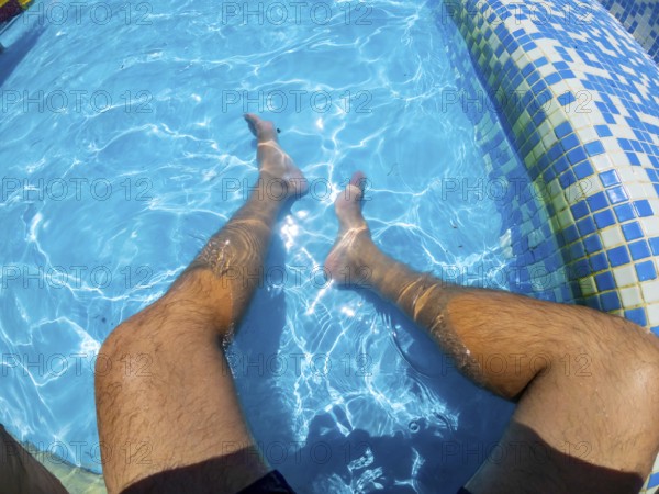 Point of view shot capturing a man relaxing in a swimming pool, legs submerged in the refreshing turquoise water under the sunny sky