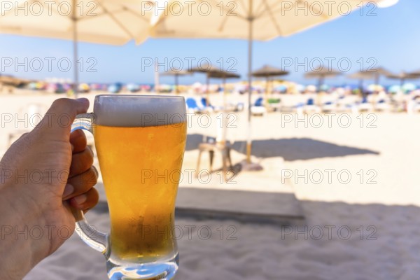 Tourist enjoying refreshing beer on sunny beach with blurred umbrellas and ocean, perfect summer vacation concept