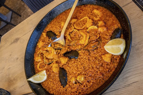 Large pan of delicious seafood paella with shrimps, mussels, squid, clams, and lemon wedges, ready to be served on a wooden table