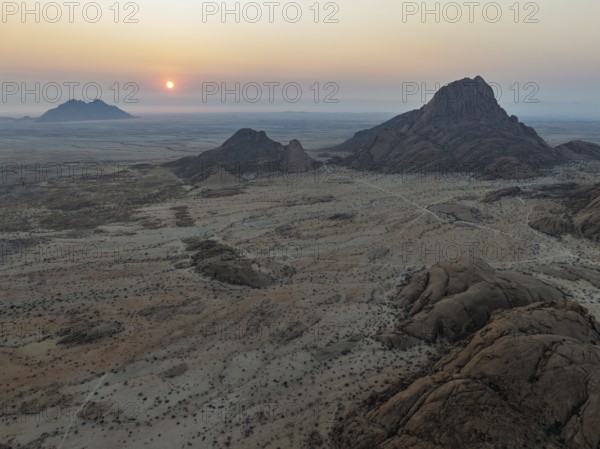 The isolated Spitzkoppe mountain (1728 m) - on the right - majestically rises above the surrounding desert plains. In the distance the coast and the Atlantic Ocean. At sunset. Aerial view. Drone shot. Damaraland, Namibia