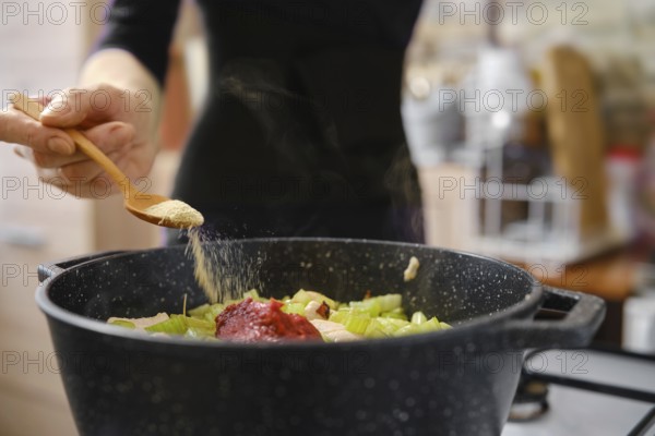 A person is adding spices to a pot filled with chopped vegetables and meat. The kitchen is warm and inviting, filled with the aroma of home-cooked food, creating a comforting atmosphere