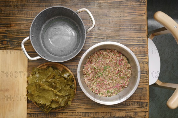 A bowl of prepared ground meat and rice mixture sits next to a plate of grape leaves ready for wrapping on a rustic wooden table