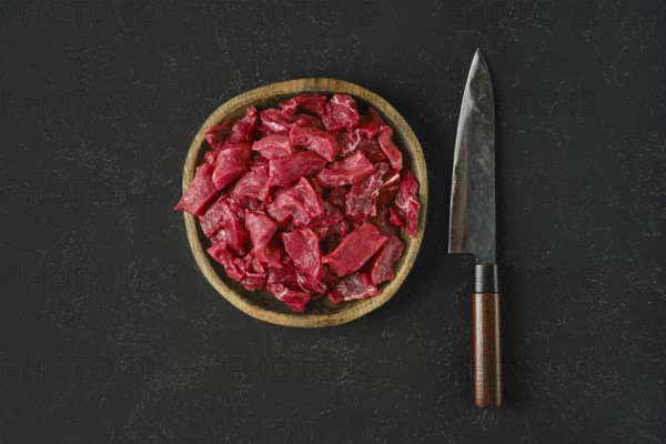 A wooden bowl holds freshly cut pieces of raw beef, showcasing a vibrant red color. Beside the bowl, a sharp knife rests on a dark textured surface, ready for preparation or cooking