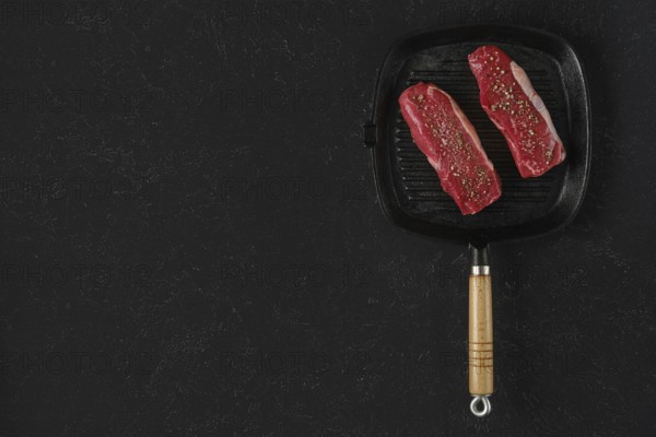 Two raw beef steaks on a preheated griddle pan, seasoned with spices, in a home kitchen setting. The black surface is for text
