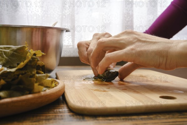 Hands delicately wrap seasoned filling in grape leaves on a wooden cutting board. Fresh ingredients and a mixing bowl are visible, showcasing the traditional culinary process of making dolma
