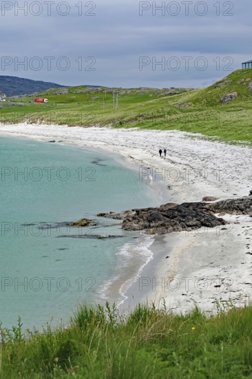 Coastal landscape with grassy areas, calm sea and walkers on a white sandy beach, Eriksay, Isle of Barra, Scotland, United Kingdom