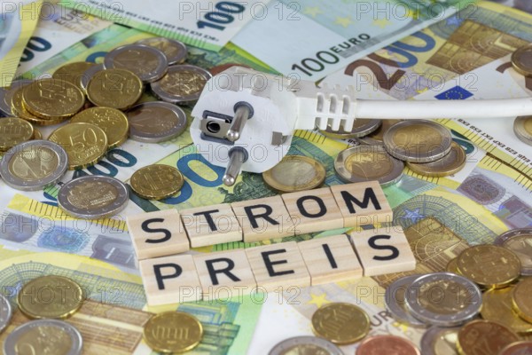 Symbolic image of electricity costs: electricity plug with euro banknotes and euro coins, with the word STROMPREIS in wooden letters in front of it
