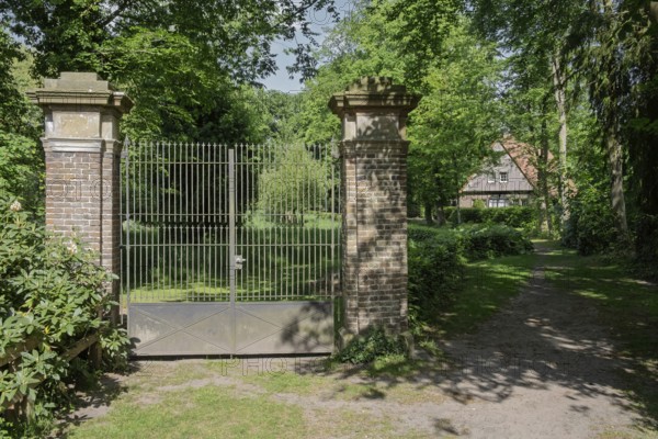 A wrought-iron gate between two brick pillars in a green, shady landscape with a building in the background, Welbergen, MÃ¼nsterland, North Rhine-Westphalia, Germany